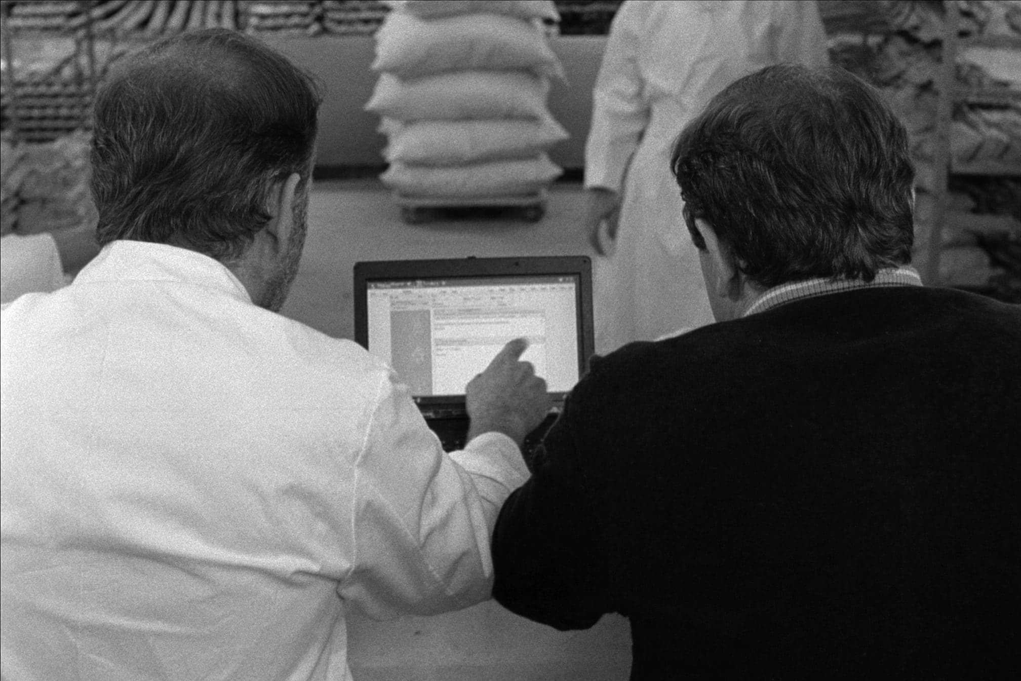 Ein Bäcker und ein Engineer sitzen nebeneinander vor einem Laptop, der Bäcker zeigt auf den Bildschirm