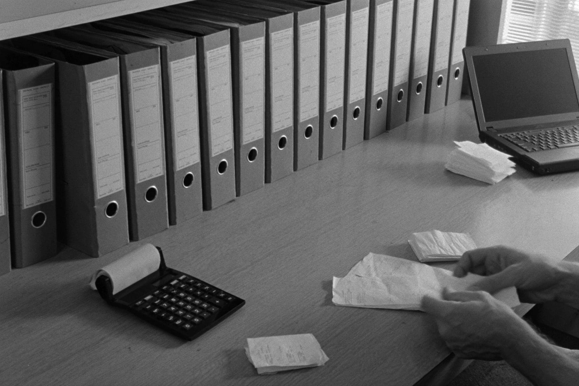 Eine Reihe grauer Bundesordner, ein Rechner mit Papierrolle, Belege und ein Laptop auf einem Bürotisch