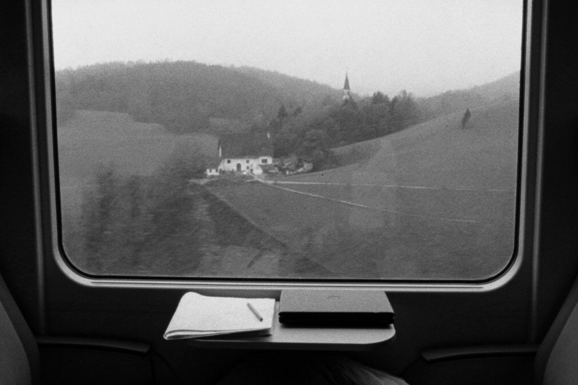 Blick aus dem Zugfenster auf Hügel, einen Bauernhof und einen Kirchturm im Nebel, auf dem Tischchen ein Notizbuch und ein Laptop